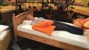 Physiotherapeutin Andy Sixtus liegt in Rückenlage auf einem Nackenstützkissen im "Wir in Bayern"-Studio in einem Bett. Ihr Arm liegt seitlich auf einem Kissen. | Bild: Wiri n Bayern