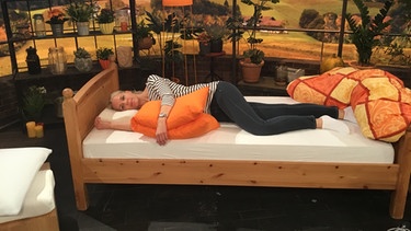 Physiotherapeutin Andy Sixtus liegt in Seitenlage auf einem Nackenstützkissen im "Wir in Bayern"-Studio in einem Bett. Ihr linker Arm liegt seitlich auf einem Kissen. | Bild: Wiri n Bayern