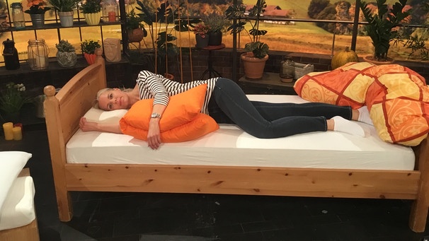 Physiotherapeutin Andy Sixtus liegt in Seitenlage auf einem Nackenstützkissen im "Wir in Bayern"-Studio in einem Bett. Ihr linker Arm und ihr linkes Bein liegen seitlich auf einem Kissen. | Bild: Wiri n Bayern