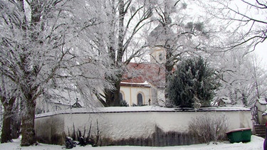 Blick auf die verschneite Pfarrkirche in Holzhausen mit Friedhof | Bild: Wir in Bayern