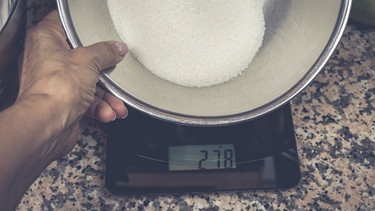 Backzutaten für einen Zitronenkuchen - Die Zuckermenge wird in einer Schüssel auf einer modernen Küchenwaage gewogen. | Bild: mauritius images / Petra Daisenberger