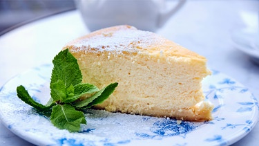 Ein Stück Käsekuchen mit Minzblättern auf einem blau-weißen Kuchenteller mit Kuchengabel auf einem Tisch mit weißer Tischdecke | Bild: mauritius images / foodcollection