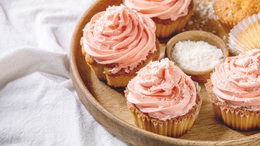 Cupcakes mit rosa Buttercreme und Kokosflocken auf einem Holztablette auf einem Tisch mit einer weißer Tischdecke | Bild: mauritius images / The Picture Pantry / Natasha Breen