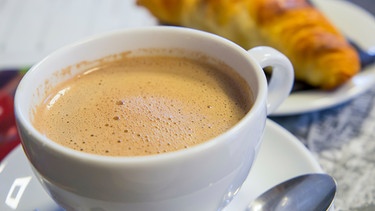 eine weiße Tasse mit Kaffee; im Hintergrund verschwommen ein Croissant auf einem weißen Teller | Bild: mauritius images / age fotostock / Ana del Castillo