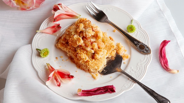 Ein Stück Apfelstreuselkuchen von oben fotografiert; der Kuchen wird auf einer Kuchengabel auf einen Kuchenteller geschoben. | Bild: 	mauritius images / Fatma Yüksel