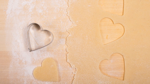von oben fotografiert: eine Arbeitsfläche aus Holz; darauf ist ein Mürbteig dünn ausgerollt und ein paar Herzen sind ausgestochen; daneben liegt ein Plätzchenausstecher in Herzform | Bild: mauritius images / Pitopia / Bernd Schmidt