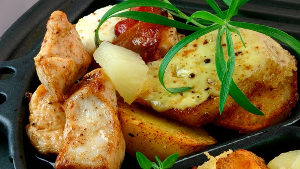 Ein Raclette-Pfännchen gefüllt mit Hühnchenbruststücken, Kartoffeln, die mit Käse überbacken sind, einem Stück Ananas, gebratenem Speck und etwas Rosmarinstängel | Bild: Wir in Bayern