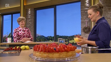 Im Vordergrund ein Erdbeer-Zitronen-Kuchen und im Hintergrund die Moderatorin Sandra Bouscarrut und die Kondormeisterin Martina Harrecker | Bild: Wir in Bayern