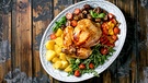 Von oben fotografiert: eine weiße Schale mit einem ganzen gegrillten Hähnchen, Kartoffeln, Tomaten und Gemüse auf einem Holztisch | Bild: mauritius images / The Picture Pantry / Natasha Breen