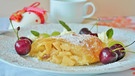 Ein Stück Apfelstrudel auf einem weißen Kuchenteller, dekoriert mit Kirschen, Minzblättern, alles ist mit Puderzucker bestäubt; im Hintergrund eine weiße Tasse und ein Osterei; | Bild: Pixabay