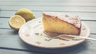 ein Stück Zitronenkuchen auf einem Blümchenteller mit einer Kuchengabel. Zwei Zitronen liegen neben dem Teller. | Bild: mauritius images / Petra Daisenberger