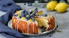 ein runder Zitronen-Blaubeer-Kuchen mit Heidelbeeren auf einem Tisch mit hellgrauer Tischdecke und zwei Zitronen im Hintergrund | Bild: mauritius images / Pitopia / Karl Allgäuer