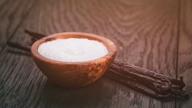 Ein Holzschälchen mit Zucker und vier Vanilleschoten daneben auf einem Holztisch | Bild: mauritius images / Vladislav Gudovskiy / Alamy