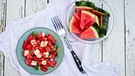 Wassermelonensalat mit Feta, Minze und Sesam von oben fotografiert, in einer hellblauen Schale; daneben eine Glasschale mit Wassermelonenstücke; alles ist auf einem weiß lasierten Holztisch | Bild: mauritius images / Westend61 / Sandra Roesch