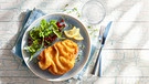 Von oben fotogrfiert: ein Teller auf einer hellblauen Stoffserviette, darauf ein Wiener Schnitzel, Zitronenscheiben und ein grüner Salat mit Granatapfelkernen, dazu ein Besteck. Alles ist in hellblau-weißen Farbtönen gehalten. | Bild: mauritius images / foodcollection / Frank Gölllner