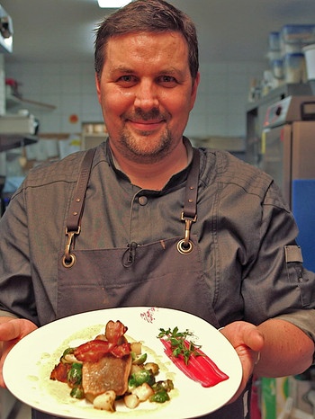 Der Koch Daniel Polzer hält einen Teller mit einem Filet von Bachsaibling auf Rosenkohl-Kartoffelgemüse mit Speckchip und Riesling-Sauce. | Bild: BR