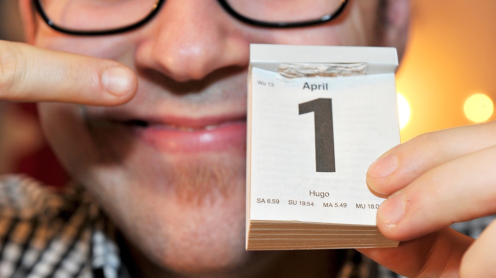 Ein Mann lacht und zeigt auf einen Kalender, dessen Datum den ersten April zeigt. | Bild: picture alliance / dpa | Felix Hörhager