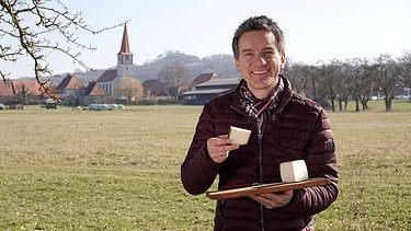 Florian Schrei hält ein Holzbrett und einem Stück Käse darauf. | Bild: BR / Kathrin Lindauer