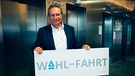 Klaus Ernst steht vor Aufzügen und hält ein Wahlfahrt Schild | Bild: BR/Salvan Joachim