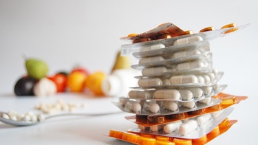 Verschiedene Medikamente aufeinandergestapelt, im Hintergrund Löffel mit Pillen + Obst/Gemüse | Bild: Pixabay