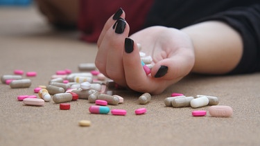 Ein Mensch liegt am Boden mit ausgestreckter Hand, darauf befinden sich jede Menge verschiedener Pillen und auf dem Boden daneben | Bild: Pixabay