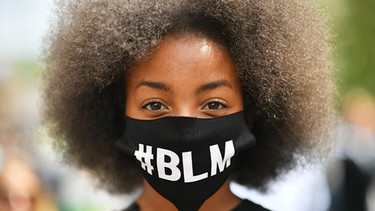 07.06.2020, Großbritannien, Bristol: Ein Demonstrantin trägt einen Stoffmaske mit dem Aufdruck «#BLM» bei einem Protest gegen Rassismus und Polizeigewalt in College Green. Weltweit drücken Menschen ihre Solidarität nach dem gewaltsamen Tod von George Floyd durch einen weißen Polizisten am 25. Mai in der US-Stadt Minneapolis aus. Foto: Ben Birchall/PA Wire/dpa +++ dpa-Bildfunk +++ | Bild: dpa-Bildfunk/Ben Birchall