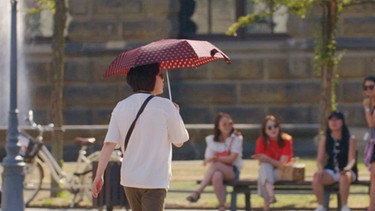 Der Regenschirm dient auch als Sonnenschutz bei der Hitzewelle. | Bild: BR/Hoferichter & Jacobs/MDR