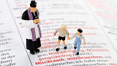 Symbolbild: Spielfiguren von Priester und Kindern auf einem aufgeschlagenen Wörterbuch über dem Wort "Missbrauch" | Bild: mauritius-images