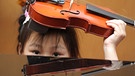 Die fünfjährige Emely blickt über ein Klavier und hält ihre Geige auf dem Kopf. Sie lernt in der Musikschule der Hofer Symphoniker Geige spielen.  | Bild: picture-alliance/dpa