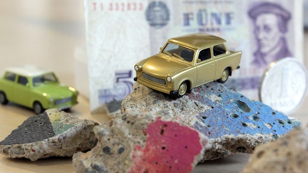 Ein Spielzeugtrabi auf einem DDR-Mauerstück und Münzen und Geldschein der DDR-Mark | Bild: BR-Studio Franken/Frank Staudenmayer