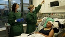 Eine Patientin liegt in einem  Krankenhausbett. Daneben stehen zwei Ärztinnen. | Bild: Bayerischer Rundfunk 2024