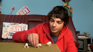 Checker Can spielt auf dem Tisch seiner Checker-Bude mit kleinen Spielzeugschweinen | Bild: Bayerischer Rundfunk 2022
