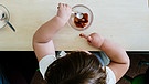 Symbolbild: Ein Kind mit offensichtlichen "Speckröllchen" von oben fotografiert, am Esstisch sitzend. Es löffelt ein Dessert. | Bild: picture-alliance/dpa