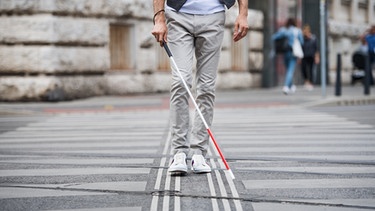 Ein blinder junger Mann überquert mithilfe eines Blindenstocks die Straße. | Bild: stock.adobe.com/Halfpoint
