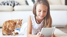 Ein Mädchen liegt mit Kopfhörern auf dem Teppich des Wohnzimmerbodens vor der Couch und blickt auf ein Tablet. Neben ihr sitzt eine rot-weiße Katze und schleckt sich die Pfote. | Bild: stock.adobe.com/Alena Ozerova
