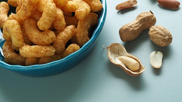 Eine Schüssel mit Erdnussflips und Erdnüsse liegen auf einem Tisch. | Bild: colourbox.com
