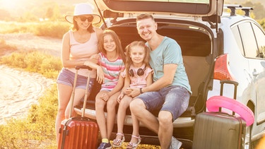 Eine glückliche Familie mit Vater, Mutter und zwei Töchtern sitzt mit ihren Koffern im Kofferaum eines Autos. | Bild: colourbox.com