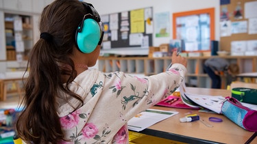 Ein Mädchen einer 2. Klasse arbeitet in der Grundschule St. Konrad im Klassenzimmer mit einem Gehörschutz an einem Arbeitsblatt. | Bild: picture-alliance/dpa