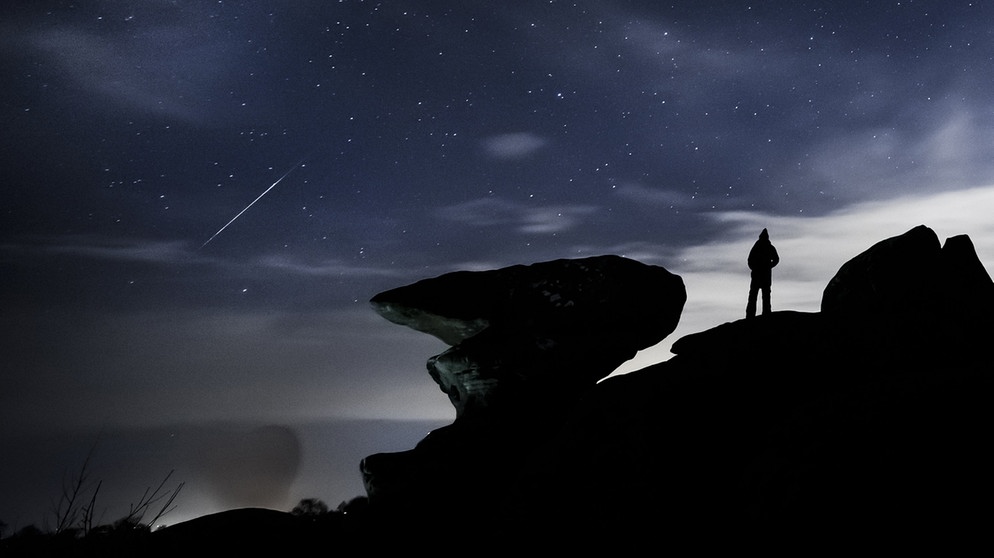 Ein Mann beobachtet im Dezember 2020 einen Meteor über der Felsformation Brimham Rocks während des Geminiden-Meteoritenschauers.  | Bild: dpa-Bildfunk/Danny Lawson