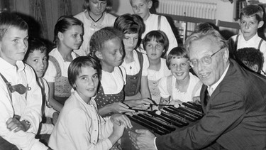 Das Foto aus dem Jahar 1964 zeigt Carl Orff mit Kindern des SOS-Kinderdorfs in Dießen am Ammersee. Der Komponist erklärt ihnen, wie man mit einem Xylophon musiziert.  | Bild: picture-alliance/dpa