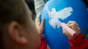 Ein Kind bemalt einen Luftballon mit Friedenstaube mit Kreide | Bild: picture-alliance/dpa
