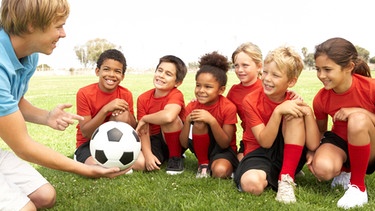 Ein jugendlicher Fußballtrainer sitzt mit Kindern auf dem Rasen und gibt ihnen Anweisungen. | Bild: colourbox.com
