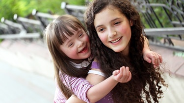 Zwei Mädchen lachen miteinander: Die große Schwester trägt die kleinere Schwester Huckepack. Das kleine Mädchen hat das Down-Syndrom.  | Bild: colourbox.com