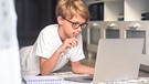 Ein Junge mit Brille liegt auf dem Wohnzimmerboden und blickt in ein großes Laptop. Daneben liegt ein Block. | Bild: stock.adobe.com/FABIO PRINCIPE