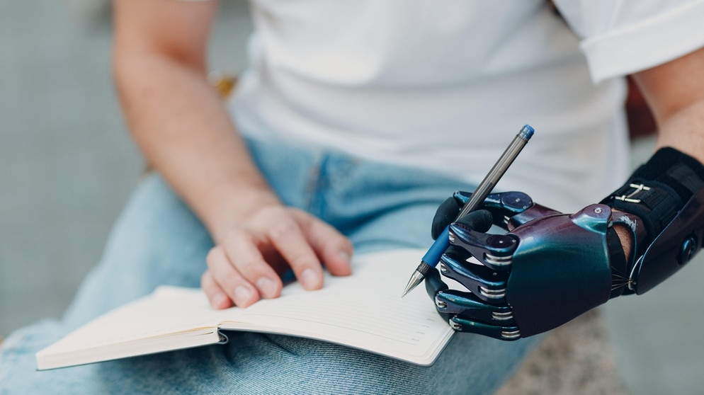 Ein junger Mann mit einer Armprothese schreibt mit seiner künstlichen linken Hand mit einem Stift in ein Schreibheft. | Bild: picture alliance / Zoonar | Max