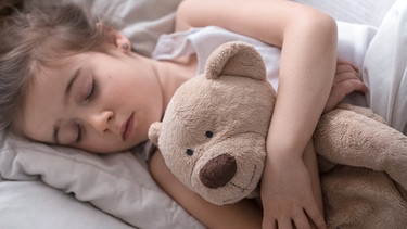 Ein Mädchen schläft mit ihrem Teddy im Arm in einem Bett. | Bild: colourbox.com
