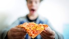 Ein Junge (verschwommen) isst ein Stück Tiefkühlpizza. | Bild: picture-alliance/dpa