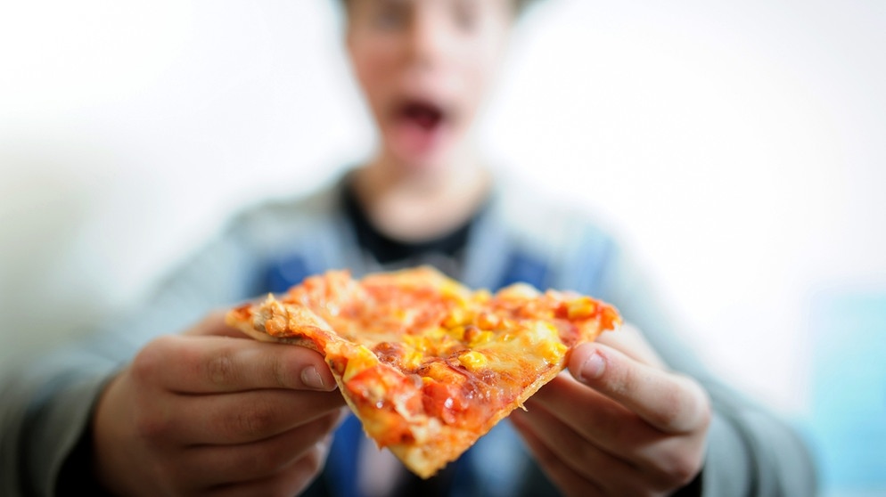 Ein Junge (verschwommen) isst ein Stück Tiefkühlpizza. | Bild: picture-alliance/dpa