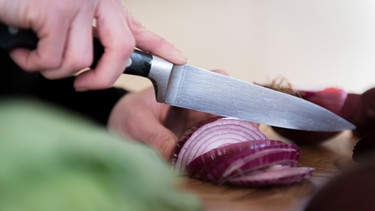 Frauenhände schneiden mit einem Messer eine rote Zwiebel. | Bild: mauritius images / TPP / Craig Holmes