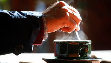 Ein Gast rührt in der Tasse an einem Tisch im Außenbereich eines Cafes: Hand in Nahaufnahme, aufsteigender Dampf über der Tasse. | Bild: dpa-Bildfunk/Jean-Francois Badias